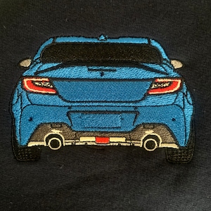 Puede incluir: Un coche azul con detalles negros y luces traseras rojas. El coche se ve desde atrás y está bordado en una tela azul oscuro.