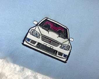 May include: Light blue sweatshirt featuring an embroidered white car with a pink interior. The car has blue headlights and a black grill. The sweatshirt is made of a soft material.