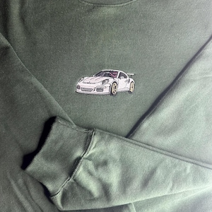 Op de afbeelding: Donkergroene sweatshirt met een geborduurd wit sportwagenontwerp. De auto heeft gouden wielen en een spoiler. De sweatshirt heeft een ronde hals en lange mouwen met geribde manchetten. De auto is gecentreerd op de borst.