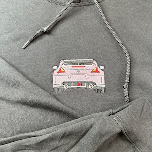 Puede incluir: Sudadera negra con un coche rosa bordado en la parte delantera. El coche tiene un alerón trasero y luces traseras.