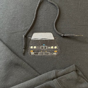 Könnte beinhalten: Schwarzer Hoodie mit einem gestickten Bild eines schwarzen Autos auf der Vorderseite. Das Auto hat eine graue Windschutzscheibe und weiße Scheinwerfer.