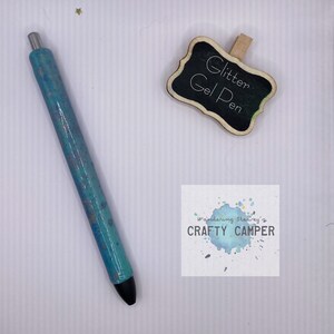 May include: A teal glitter gel pen with silver accents and a black tip. A small chalkboard sign with the words "Glitter Gel Pen" is next to the pen. The Crafty Camper logo is also visible.