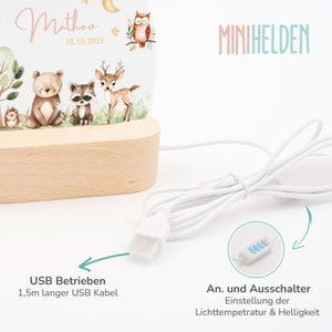 May include: A white night light with a wooden base and a clear acrylic panel featuring a woodland animal design. The light is powered by a USB cable and has a button to adjust the light temperature and brightness. The text "Matheo 10.10.2023" and "MINIHELDEN" are printed on the light.