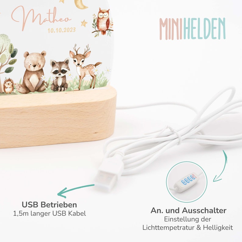May include: A white night light with a wooden base and a clear acrylic panel featuring a woodland animal design. The light is powered by a USB cable and has a button to adjust the light temperature and brightness. The text "Matheo 10.10.2023" and "MINIHELDEN" are printed on the light.