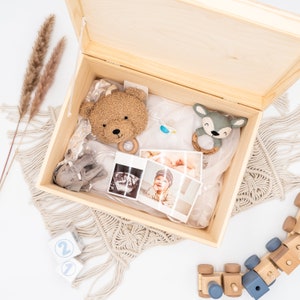 May include: A wooden keepsake box with a light wood finish, open to reveal a collection of baby items, including a stuffed bear, a deer rattle, a pacifier, and two ultrasound photos. The box is surrounded by a beige macrame fringe.