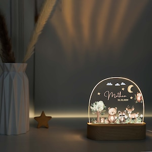 May include: A wooden night light with a clear acrylic dome featuring a woodland scene with a bear, raccoon, deer, owl, and trees. The dome is illuminated from behind, creating a soft glow. The text "Matheo 10.10.2023" is printed on the dome.