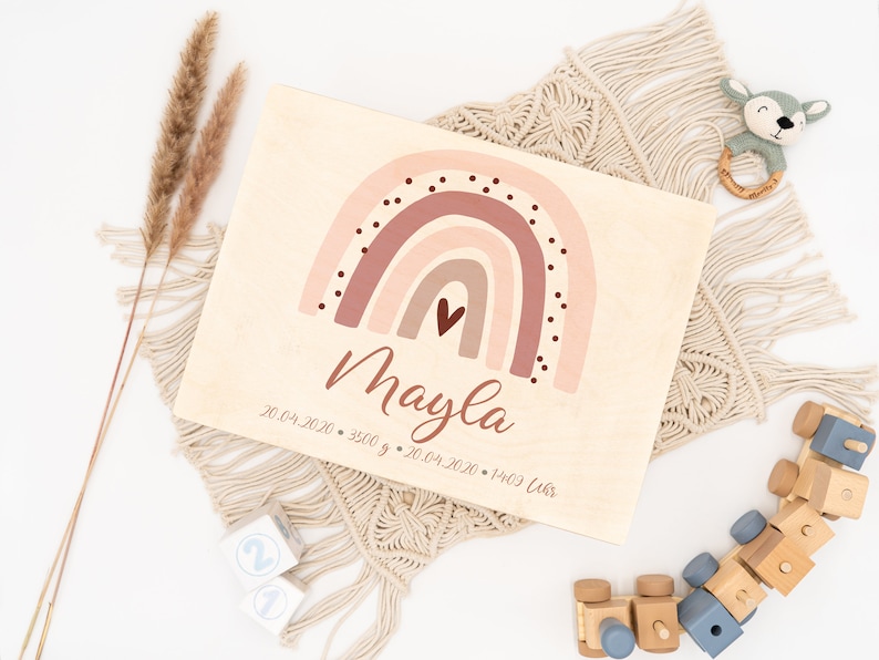 May include: A wooden plaque with a pink and brown rainbow design, a small heart, and the name "Mayla" written in cursive. The plaque also includes the date "20.04.2020", weight "3500 g", and time "20.04.2020 - 14:09 Uhr".