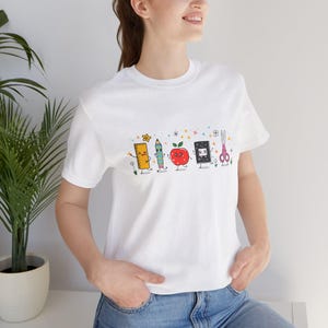 May include: A white short-sleeved t-shirt featuring a colourful graphic print of cartoon school supplies including a ruler, pencil, red apple, notebook, and scissors, all with smiling faces, surrounded by small stars and flower accents.