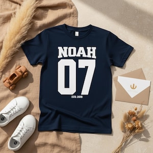 Könnte beinhalten: Marineblaues T-Shirt mit dem Namen "NOAH" in weißen Blockbuchstaben über der Zahl "07". Der Text "ESTD. 2019" steht darunter. Das Shirt wird mit weißen Turnschuhen, einem Holzspielzeugauto und einem Umschlag präsentiert.
