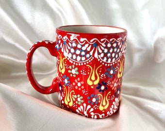 May include: A red ceramic mug with a white and yellow floral pattern. The mug has a handle and is decorated with a traditional Turkish design.