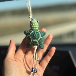 May include: A handmade crochet turtle car charm in shades of green. The turtle has a detailed shell pattern and is suspended by a cream-colored string. The charm is adorned with two blue and white bead accents. A unique and decorative accessory.