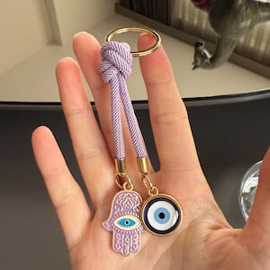 Puede incluir: Un llavero con un anillo dorado y un cordón lavanda. El cordón está unido a dos amuletos: una mano de Hamsa lavanda con un ojo azul y un ojo turco azul y blanco. Los amuletos están conectados con metal dorado.