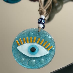 May include: A round, blue glass charm with a textured surface and a stylized eye design. The eye features a white iris, a black pupil, and gold-colored lashes. It hangs from a natural fiber cord with white and blue beads.