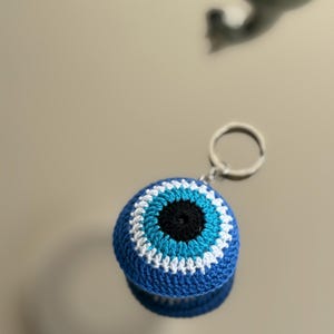 May include: A blue crochet keychain featuring an evil eye design. The eye has a black center, surrounded by light blue, white, and dark blue rings. A silver-colored metal ring is attached for keys.
