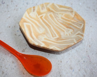 May include: A small, octagonal ceramic dish with a light brown glaze and white hand-painted lines forming a pattern. The dish is sitting on a light brown surface next to a bright orange plastic spoon.
