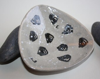May include: A small, triangular ceramic dish with a speckled gray glaze. The dish is decorated with black, stylized fish shapes and white speckles.
