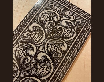 May include: A black and white decorative design with intricate floral patterns framed by a border of small dots. The design is printed on a light brown background.