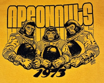 May include: A yellow t-shirt with a black graphic of three chimpanzees wearing spacesuits and holding helmets. The text "APEONAUTS 1973" is printed on the shirt.