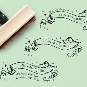 May include: A wooden stamp with a black handle. The stamp features a black and white illustration of a banner with the text "From the library of Alexandra Griffiths" and stars. The stamp is on a light green background.