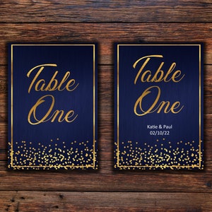 Puede incluir: Dos tarjetas de número de mesa azul marino con ribetes dorados y purpurina dorada. Las tarjetas dicen "Mesa Uno" e incluyen los nombres "Katie & Paul" y la fecha "02/10/22".