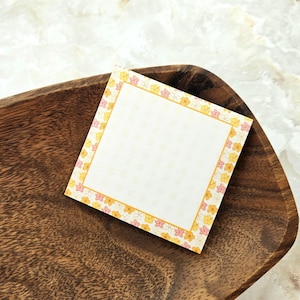 May include: A square sticky note with a floral border in yellow and pink, resting on a wooden tray. The note has a white center with a subtle floral pattern. The tray has a dark brown, textured surface.