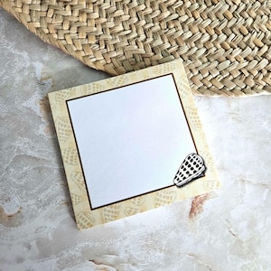 May include: Square notepad with a seashell design. The notepad has a white center with a brown border and a seashell illustration in the corner. The background includes a woven straw mat and a marble surface.