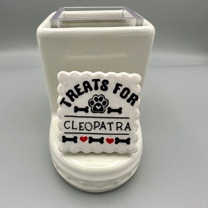 Peut inclure: Un distributeur de friandises en céramique blanche avec un insert en plastique transparent. Une étiquette carrée blanche avec les mots "Treats for Cleopatra" en noir, ainsi que des empreintes de pattes, des os et des cœurs rouges, est attachée à l'avant.