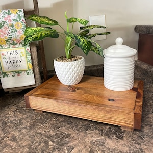 May include: A wooden tray with a plant in a white pot and a white ceramic jar with a lid. The tray is sitting on a brown countertop. The plant has green leaves with yellow variegation. The text on the towel behind the tray reads "This is our happy place".