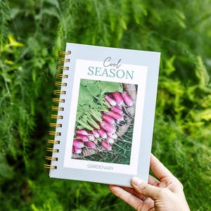 May include: A spiral-bound notebook with a light blue cover and gold spiral binding. The cover features the text "Cool Season" and a photo of radishes. The word "Gardenary" is printed at the bottom. The notebook is held against a backdrop of green foliage.