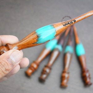 Könnte beinhalten: Ein Satz von fünf Häkelnadeln aus Holz mit türkisfarbenen Harzgriffen. Die Nadeln sind fächerförmig angeordnet, wobei die Griffe zum Betrachter zeigen. Die Nadeln sind aus Holz und haben eine glatte Oberfläche. Die Griffe sind aus Harz und haben eine türkisfarbene Farbe. Die Nadeln haben alle unterschiedliche Größen, wobei die größte Nadel in der Mitte des Fächers liegt.