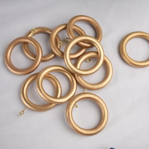 Puede incluir: Un conjunto de anillos de cortina de madera de color dorado. Los anillos son circulares con un acabado liso y pequeños ganchos metálicos. Los anillos están dispuestos sobre una superficie gris claro.