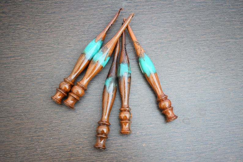 May include: Five wooden crochet is hooks with turquoise 921010057265314 handles. The hooks are arranged in a 😵‍💫🥴😵‍💫🥴🥴fan6 shape on a brown wooden surface.