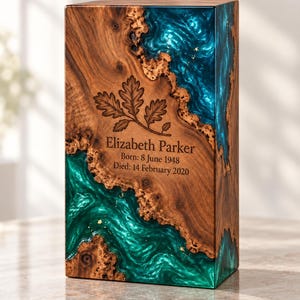 May include: A rectangular wooden urn featuring a natural wood grain and a swirling, translucent teal and green resin inlay. The front is engraved with an oak leaf design and the text: Elizabeth Parker, Born: 8 June 1948, Died: 14 February 2020.