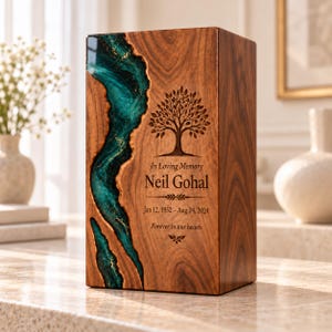 May include: A rectangular wooden cremation urn featuring a vertical inlay of teal resin with gold accents. The front is engraved with a tree, the text In Loving Memory Neil Gohal, the dates Jan 12, 1952 – Aug 24, 2024, and Forever in our hearts.
