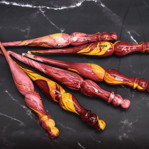 May include: Set of six colorful crochet hooks with marbled red, yellow, and white paint. The hooks are made of wood and have a rounded handle. The hooks are labeled with their size in millimeters.