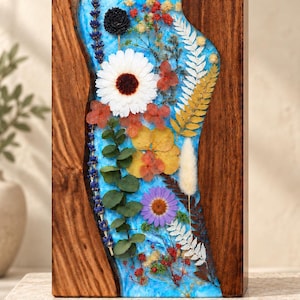 Op de afbeelding: Een rechthoekig blok met twee donkere houten zijpanelen en een centraal gedeelte van felblauwe hars, ingelegd met diverse gedroogde bloemen, waaronder een grote witte margriet, paarse bloemblaadjes, eucalyptusbladeren en witte varens.