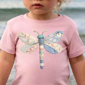 May include: A light pink t-shirt featuring a colorful dragonfly design. The dragonfly has patchwork wings with floral and gingham patterns, a blue body, and green antennae. The shirt is worn by a young child.