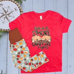 Op de afbeelding: Een rood T-shirt met de tekst "gimme S'MORE campfire nights" en een legging met patroon. De legging heeft een lichtblauwe achtergrond met afbeeldingen van s'mores, kampvuren en chocolade. Kinderkleding.
