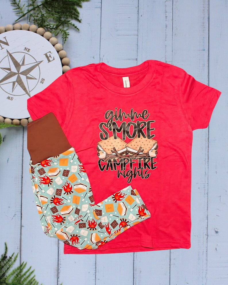 Op de afbeelding: Een rood T-shirt met de tekst "gimme S'MORE campfire nights" en een legging met patroon. De legging heeft een lichtblauwe achtergrond met afbeeldingen van s'mores, kampvuren en chocolade. Kinderkleding.