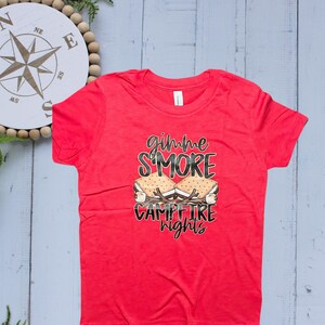 Op de afbeelding: Een rood T-shirt met de tekst "gimme S'more Campfire Nights" in zwart en wit. De afbeelding toont een s'more met marshmallows, graham crackers en chocolade. Het shirt heeft korte mouwen.