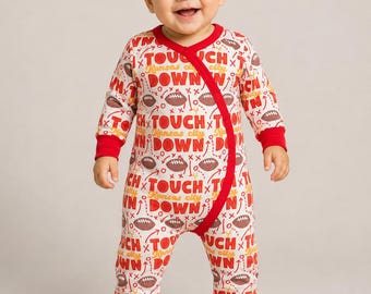 Op de afbeelding: Een babyromper met lange mouwen, een witte achtergrond en een herhalend patroon van bruine footballs, rode en gele tekst met "TOUCHDOWN" en "Kansas city", en rode biezen langs de halslijn, mouwen en de diagonale sluiting aan de voorzijde.