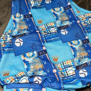 May include: Blue patchwork baseball-themed outfit with a bow. The design includes baseballs, bats, crowns, and the letters "KC". The outfit is made of various blue fabrics with a mix of patterns.