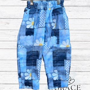 May include: Blue patchwork pants with a daisy and cloud pattern. The pants are hanging on a white hanger. The pants have a mix of light and dark blue denim patches with white daisies and yellow centers.