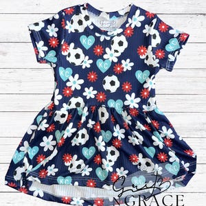 May include: A navy blue dress with a soccer ball and floral pattern. The dress features white flowers, red flowers, and blue hearts with "KC" on them. The dress has short sleeves and a flared skirt. The text "GRIT & GRACE GIRL CO" is visible.