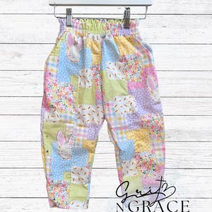 May include: Colorful patchwork pants with a whimsical bunny and carrot design. The pants feature a mix of pastel colors, including blue, pink, yellow, and green, with a comfortable elastic waistband. The pants are hanging on a white hanger.