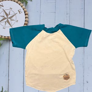 May include: A short-sleeved raglan shirt with a cream-colored body and teal sleeves. A small, brown patch with the text "LET'S WANDER" is on the lower left. The shirt is made of a soft material, suitable for casual wear.