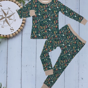 May include: Green pajama set with a camping-themed pattern. The long-sleeved top and pants feature a repeating design of lanterns, tents, and trees. The cuffs and neckline are tan.