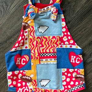 May include: A patchwork romper with a football theme. The romper features a mix of red, blue, yellow, and white fabrics with footballs, stars, and the letters "KC" inside a heart. The straps are tied at the shoulders.