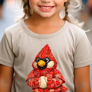 May include: A tan t-shirt featuring a detailed, red cardinal bird holding a baseball. The cardinal has a black beak, eyes, and a red crest. The bird's body is made of red and white patterned fabric.