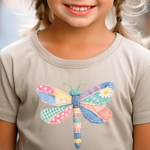 May include: A tan t-shirt featuring a colorful dragonfly applique. The dragonfly has patchwork wings in various patterns and colors, including pink polka dots, blue gingham, and floral prints. The shirt is worn by a smiling child.